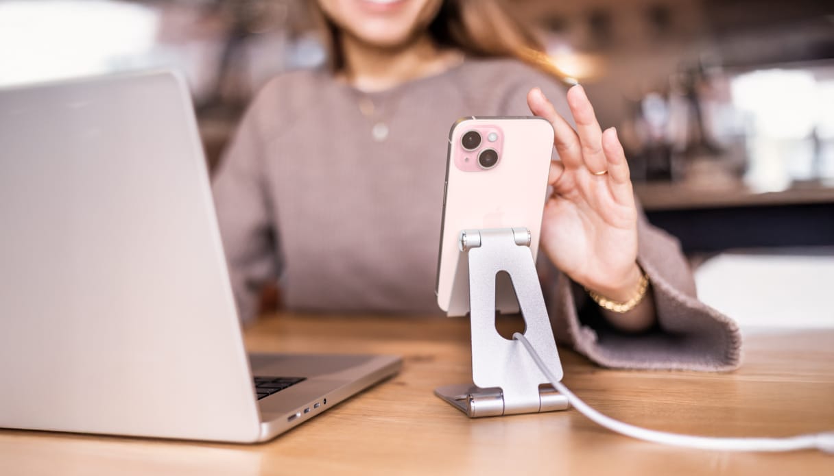 een vrouw bedient haar telefoon die in een telefoonstandaard staat terwijl hij wordt opgeladen. Op de tafel staat ook haar laptop.