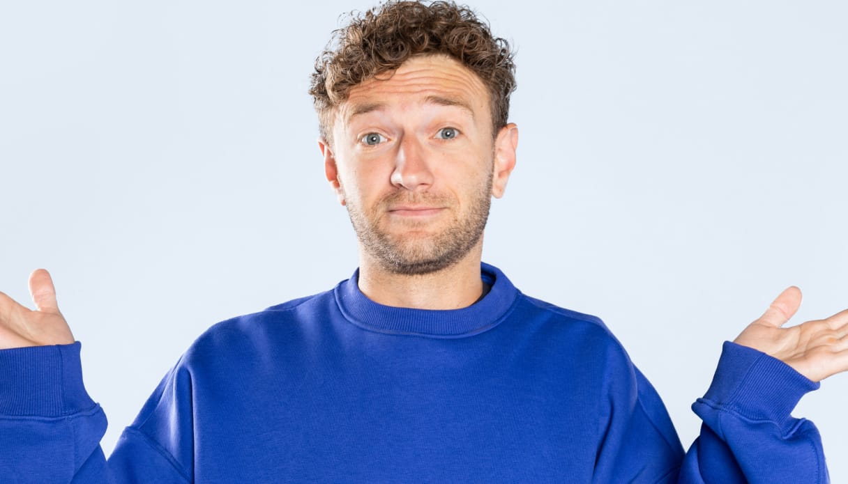 Een jongeman met krulletjes houdt vragend zijn handen omhoog. Hij draagt een blauwe trui en de achtergrond is lichtblauw.