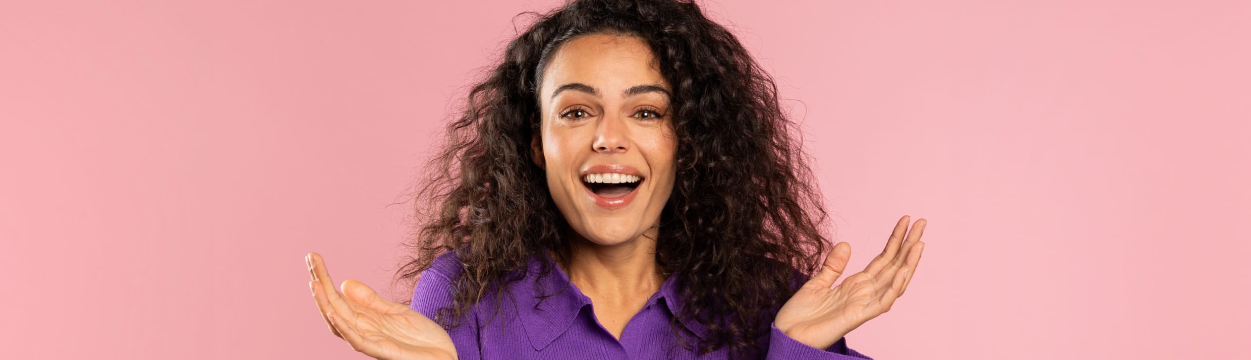 Een opgewekte vrouw kijkt recht in de lens en houd haar handen vragend omhoog. Ze heeft een bos met donkere krullen, vriendelijke ogen en de achtergrond is lichtroze van kleur.