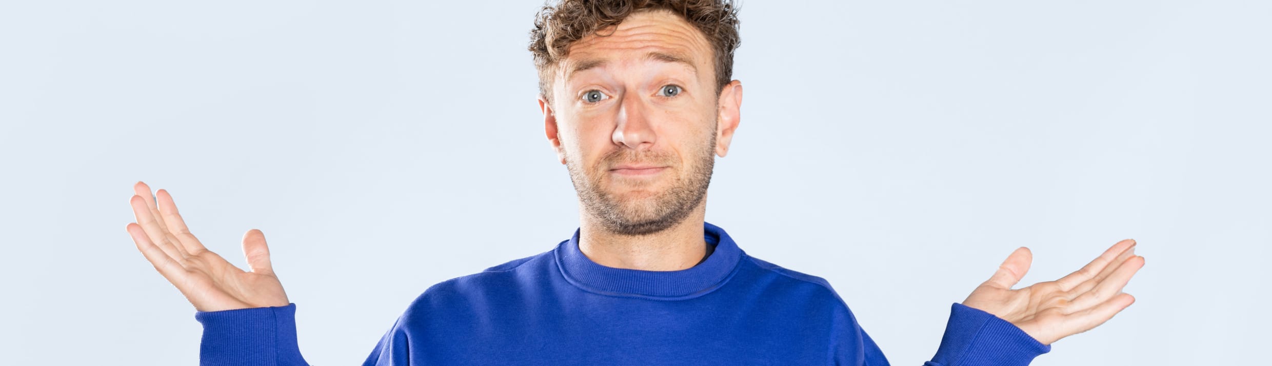 Een jongeman met een lichte huidskleur en donkerblond krullend haar heeft een vragend gezicht en houdt zijn handen vragend in de lucht. Zijn trui is blauw en de achtergrond lichtblauw.