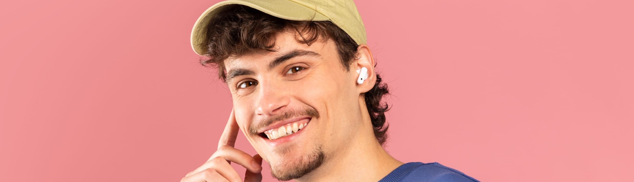 Man met lichte huid, donkere krullen luistert vrolijk naar iets op zijn draadloze witte oordopjes. Hij heeft een lichtgele pet, blauwe trui op een zachtroze achtergrond. 