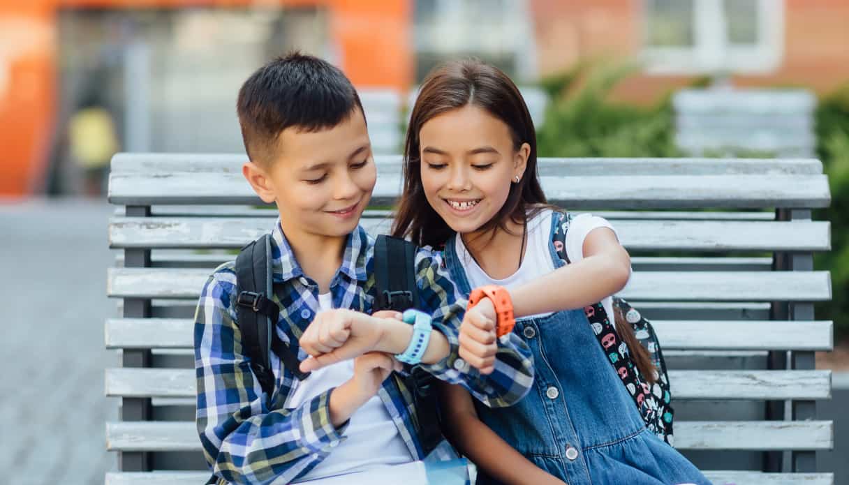 Je ziet twee kinderen op een houten bankje met elkaar kletsen en naar hun smartwatches kijken. Het jongetje links heeft een lichtblauw horloge met siliconen band, het meisje rechts oranje.