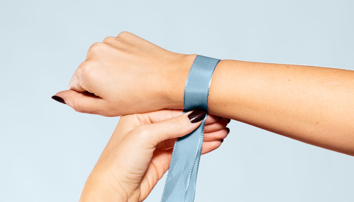 Een hand met een blauw lint om de pols waarmee de polsomtrek wordt opgemeten, de achtergrond is lichtblauw.
