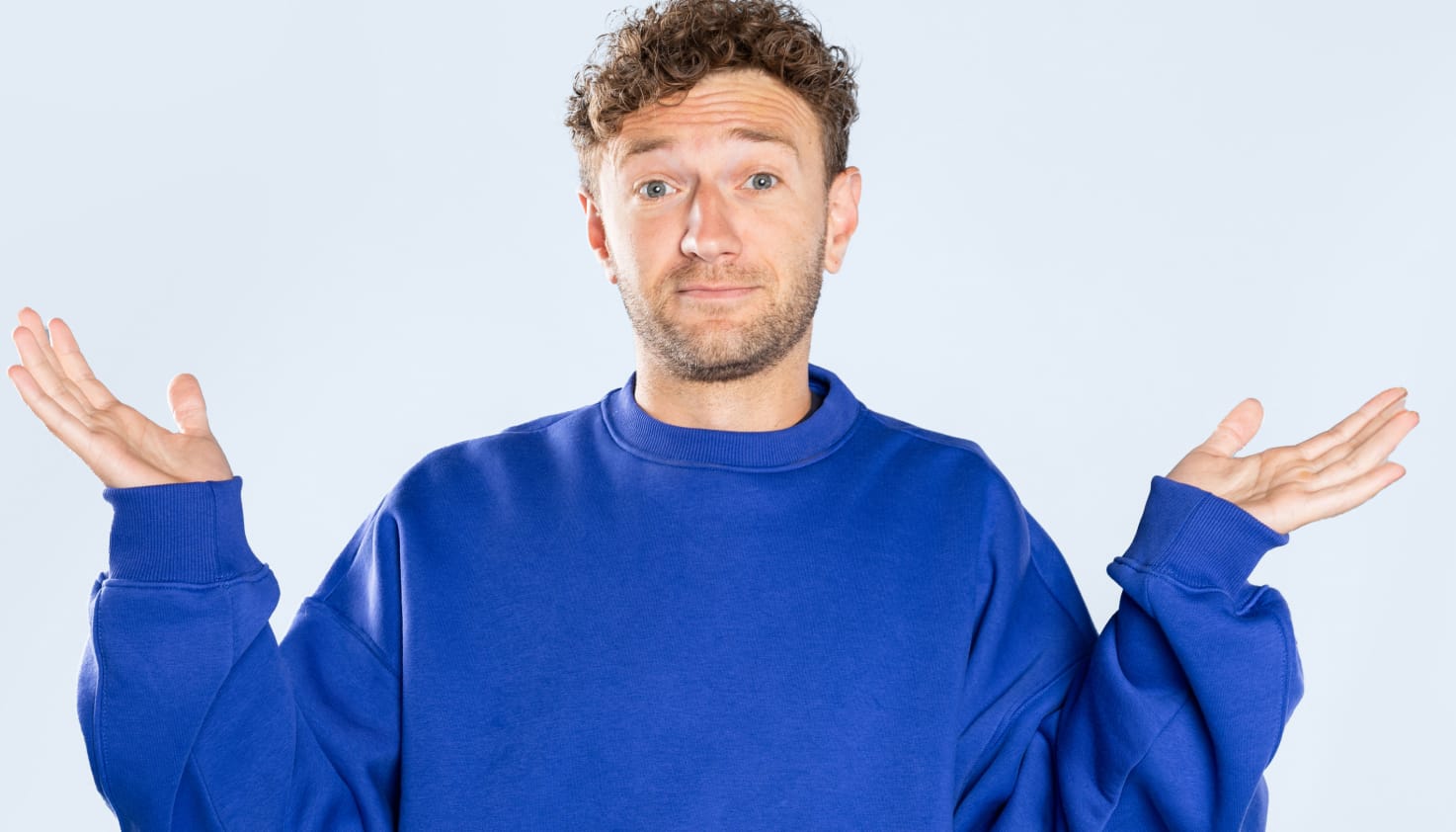 Een jongeman met krulletjes houdt vragend zijn handen omhoog. Hij draagt een blauwe trui en de achtergrond is lichtblauw.