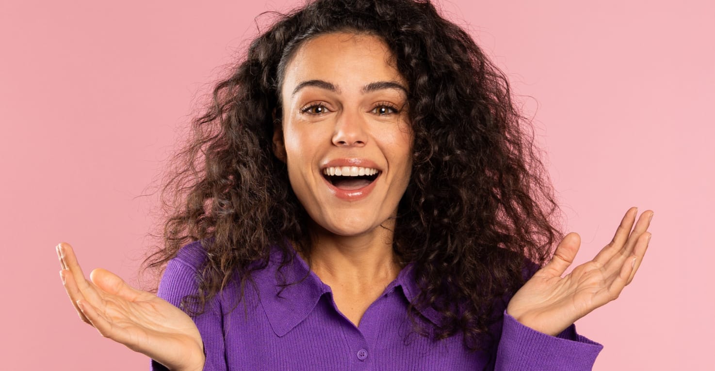 Een opgewekte vrouw kijkt recht in de lens en houd haar handen vragend omhoog. Ze heeft een bos met donkere krullen, vriendelijke ogen en de achtergrond is lichtroze van kleur.