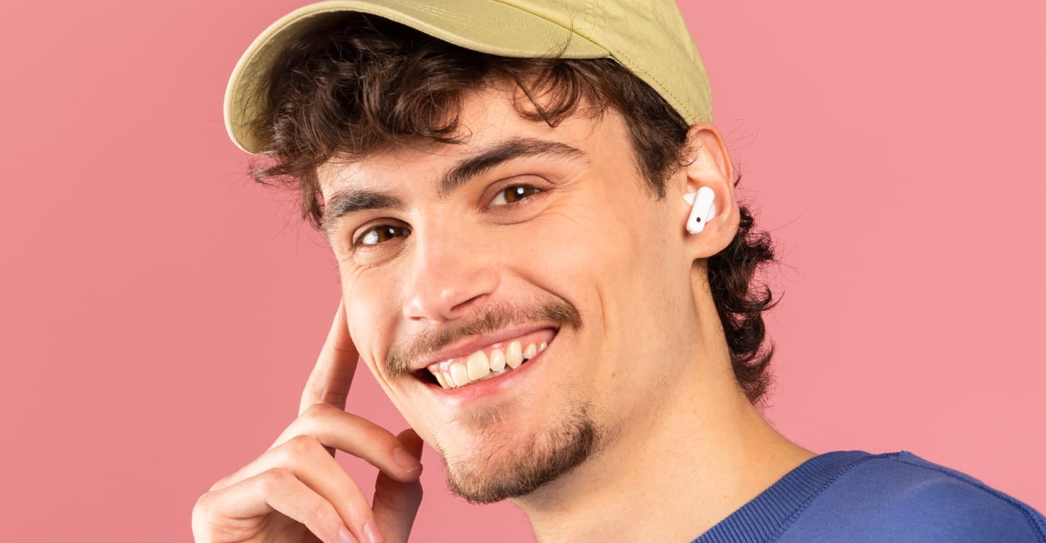 Man met lichte huid, donkere krullen luistert vrolijk naar iets op zijn draadloze witte oordopjes. Hij heeft een lichtgele pet, blauwe trui op een zachtroze achtergrond. 