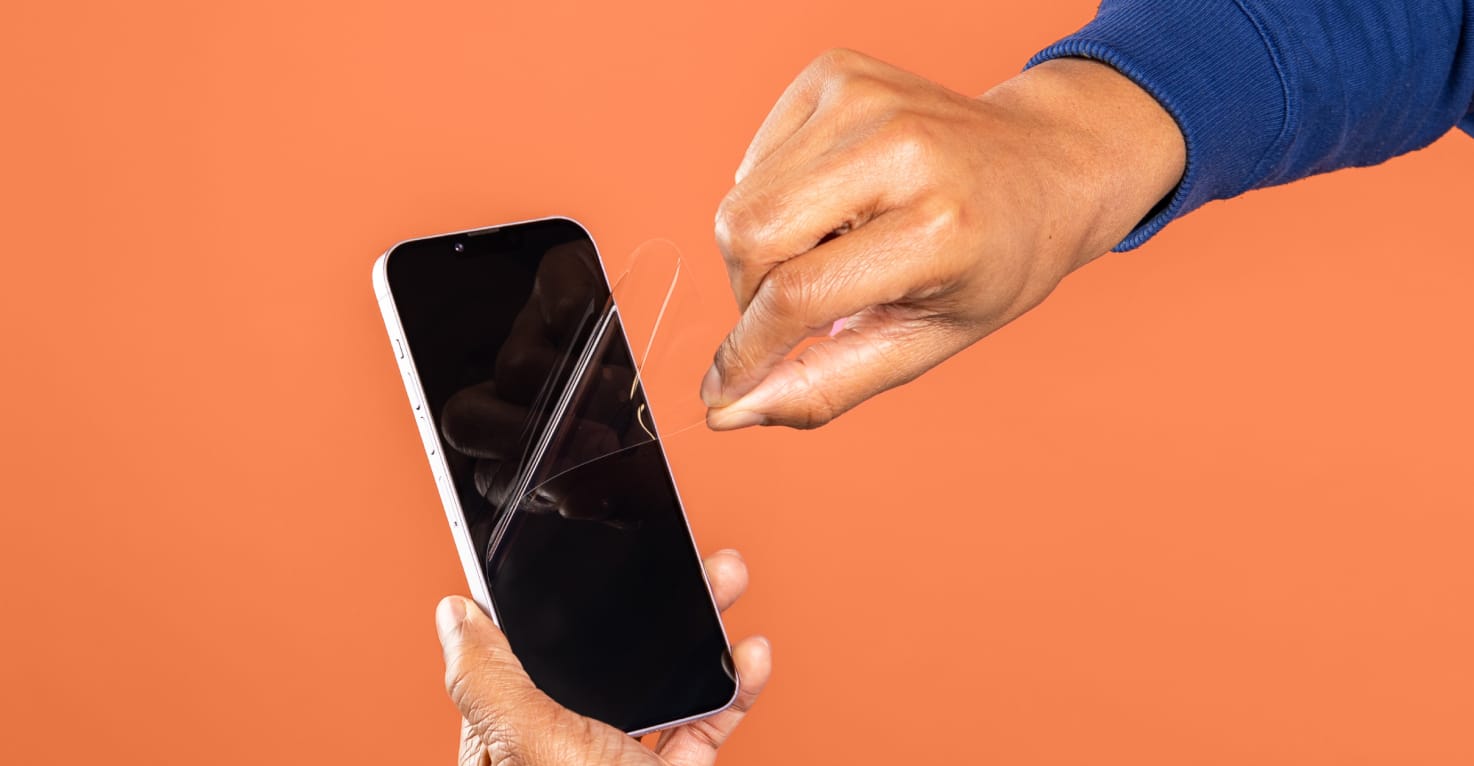 Je ziet een hand met een blauwe mauw een screenprotector verwijderen van een smartphone. De achtergrond is oranje.