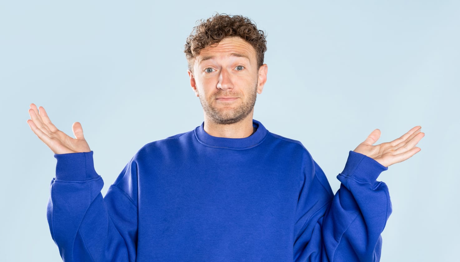 Een man in een kobalt blauwe trui steekt vragend zijn handen omhoog.