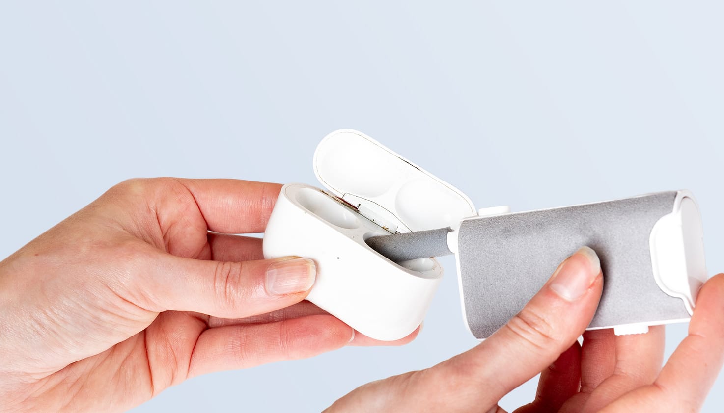 Je ziet twee handen een AirPods oplaadcase schoonmaken met een AirPod schoonmaak toolkit. Er gaat een borsteltje in de ruimte van een AirPod, de achtergrond is lichtblauw.