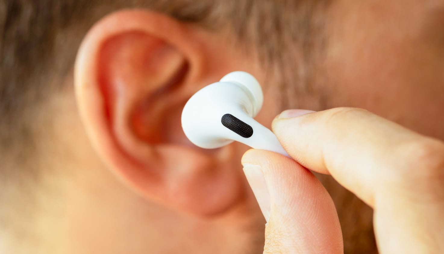 Je ziet een ingezoomd zijaanzicht van een mannenhoofd en zijn oor met donkerkleurig haar en lichte huidskleur. Hij houdt een Apple AirPod Pro voor zijn oren, deze is in focus en zijn oor is wat onscherp.