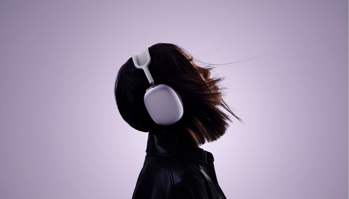 Een stijlvolle foto van een onderbelichtte dame en profil, met paarse AirPods Max op haar hoofd en haar haren waaien voor haar gezicht. De achtergrond is paars en wordt donkerder langs de randen als een vignette.