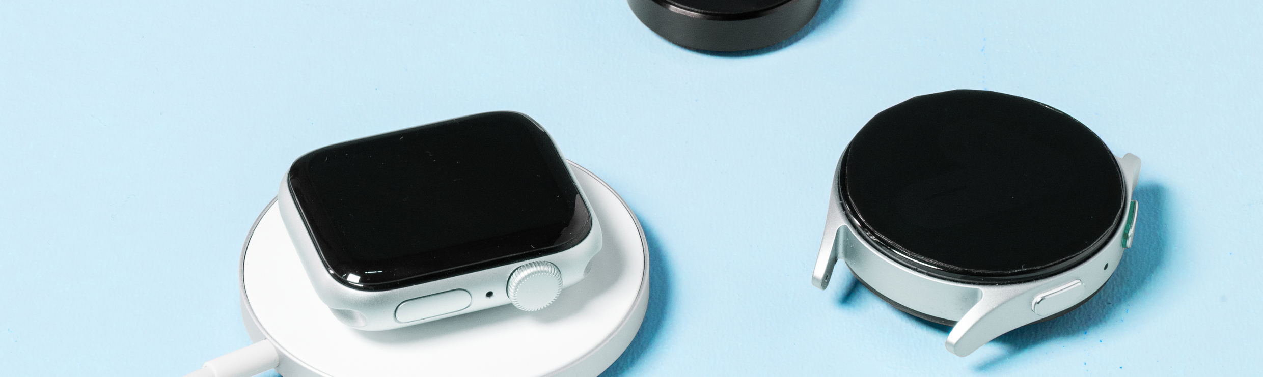 Een Apple Watch op een draadloze oplader, daarnaast ligt een Samsung Watch en een losse smartwatch oplader